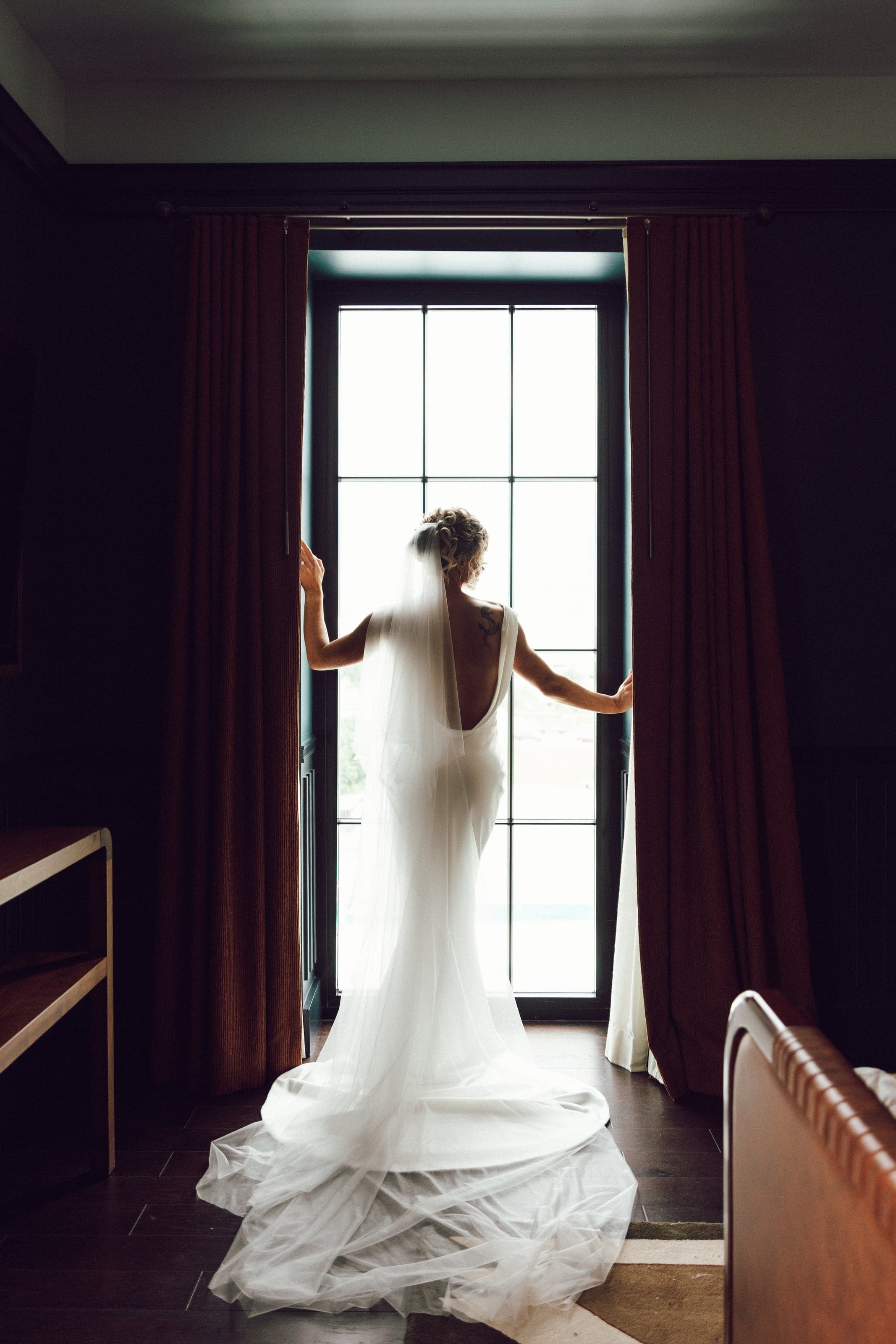 Bride alone at a tall window, veil trailing on dark floorboards